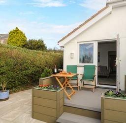 A patio with two green chairs and a wooden table with wine glasses outside a white house at Glenhaven in Trewarmett near Tintagel