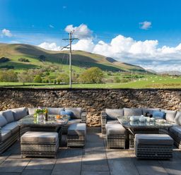 An outdoor seating area with cushioned wicker sofas and tables set on a stone patio with hills and fields in the background at Oakdene Country House in Sedbergh