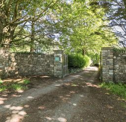 An open gate with stone walls leading to a driveway lined with trees and bushes at Culdrose Manor in Helston