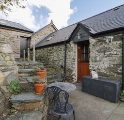 An outdoor stone patio with a metal table and chairs potted plants stone steps and a cat on a black storage box at Nyth Y Wennol in Llanfachraeth