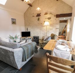 A living room with a grey sofa a wooden dining table with two chairs a green armchair a TV on a stand and a stone wall at Nyth Y Wennol in Llanfachraeth