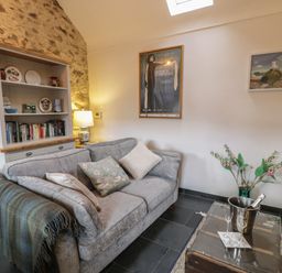 A living room with a grey sofa cushions a throw blanket a bookshelf a table with champagne and glasses flowers and framed pictures on the wall at Nyth Y Wennol in Llanfachraeth