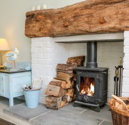 A wood burning stove with firewood stacked beside it and a blue cabinet with a lamp and decorative birds in a room at 3 Elm Cottage in Carhampton