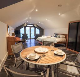 A room with a round dining table set for two, a bed, two chairs facing a window, and a wardrobe at The Boat House at Louper Weir in Bowness-On-Windermere