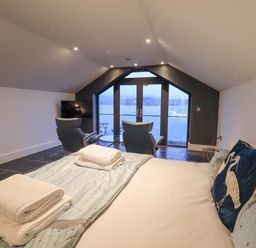 A bedroom with a bed pillows and towels two chairs a television and glass doors overlooking water at The Boat House at Louper Weir Bowness-On-Windermere