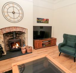 A living room with a fireplace wood stove a large clock above and a television on a wooden stand at The Old Sweet Shop in Alnwick
