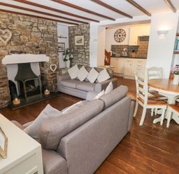 A living room with stone fireplace grey sofas a wooden dining table and kitchen area at Inglenook Cottage in Broad Haven