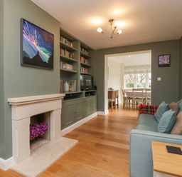 A living room with a fireplace a built in bookshelf a blue sofa and a dining room visible through a doorway at 3 Painters Field Quenington Near Cirencester