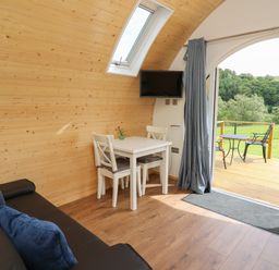 An interior room with wooden walls a small white dining table with two chairs a wall-mounted TV a black sofa with blue cushions and an open door leading to a deck with outdoor furniture and green landscape visible at Seren in Llandrindod Wells