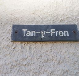 A cracked black nameplate with white letters mounted on a textured white wall at Tanyfron Holiday Studio in Bryngwran near Gwalchmai