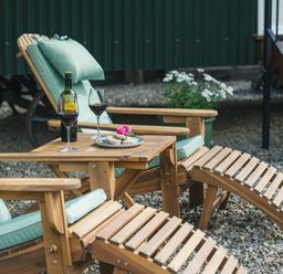Wooden lounge chairs with light green cushions and a small table holding a bottle of wine wine glasses and a plate with food on gravel at Onnen Oakford near Aberaeron