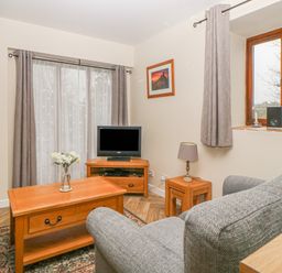 A living room with a gray sofa wooden coffee table with flowers television on a wooden stand and a window at Hilly Field Barn in Yelverton
