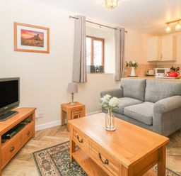 A living area with a gray sofa wooden coffee table and TV stand next to a kitchen at Hilly Field Barn in Yelverton