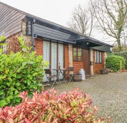 A wooden cottage with two chairs and a table outside surrounded by bushes at Hilly Field Barn in Yelverton