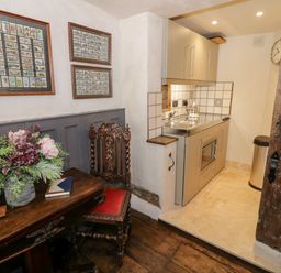 A small kitchen area with cabinets and sink next to a wooden table with flowers and a carved wooden chair at The Old Paper Shop in Tewkesbury