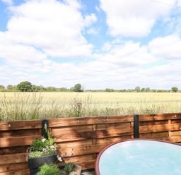 A fenced outdoor area with a hot tub and potted plants overlooking a field under a partly cloudy sky at Cobblers Barn in Repps With Bastwick near Potter Heigham