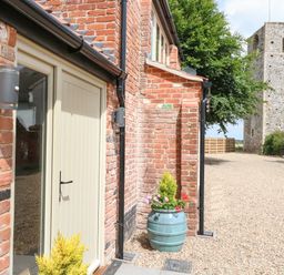 An exterior view of a brick building with a beige door and two potted plants near the entrance with a gravel driveway and a stone tower in the background at Cobblers Barn in Repps With Bastwick near Potter Heigham