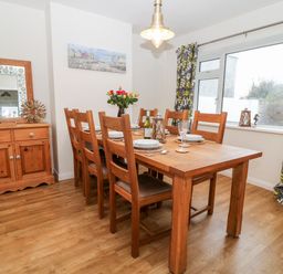 A dining room with wooden table and chairs a sideboard with a mirror and decorative items and a large window with patterned curtains at Cranberry in Bude