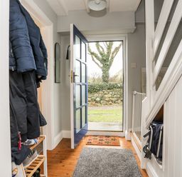 An entry hallway with coats hanging on a rack a shoe rack a blue door open to outside a tree outside and a stairway with a baby gate at The Granary - Rhoscolyn in Rhoscolyn