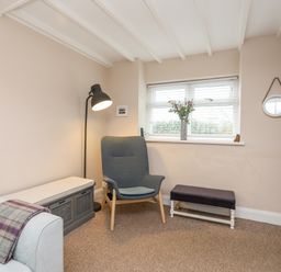 A sitting room with a gray armchair a floor lamp a window with flowers on the sill and a sofa with a blanket at The Granary - Rhoscolyn in Rhoscolyn