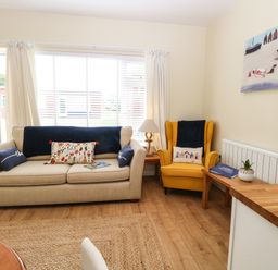 A living room with a beige sofa and yellow armchair near a window with white curtains at The Cranny in Mundesley