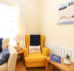 A corner with a yellow armchair a beige sofa side table with a lamp another side table with a plant and book and a painting on the wall at The Cranny in Mundesley