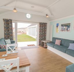 A living room with blue sofas and patterned cushions a wooden dining table white chairs and framed pictures on a light blue wall at Shore Thing 280 in Wilsthorpe