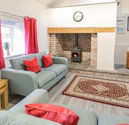 A living room with grey sofas and red cushions a brick fireplace with a wood stove a red patterned rug and a television at Crud yr Awel Braichmelyn near Bethesda
