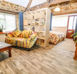 A studio room with a colorful sofa a wooden coffee table a bed separated by a cubed bookshelf and stone walls at The Forge at Smithy Cottage in Bloxham