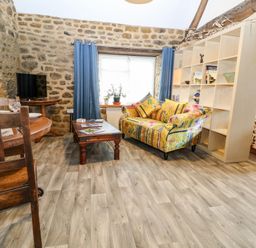 A living room with a wooden dining table and chairs a colorful sofa a wooden coffee table a shelving unit and blue curtains at The Forge at Smithy Cottage in Bloxham
