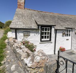 Exterior view of a white cottage with stone roof next to a stone wall and a gravel path at Harbour Cottage in Boscastle