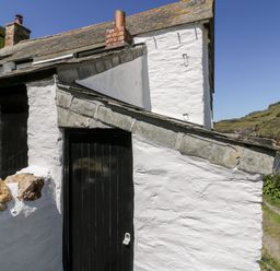A white stone cottage with black doors and a sloped stone roof near a rocky hillside and water at Harbour Cottage in Boscastle