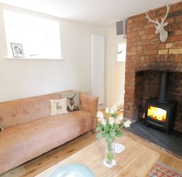 A living room with a brown sofa with animal print cushions a wooden coffee table with flowers and a brick fireplace with a wood stove at Prospect Cottage in Malvern Wells near Great Malvern