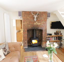 A living room with a brick fireplace a wood stove a television a bookshelf a beige sofa with deer pillows a wooden coffee table with flowers and a staircase at Prospect Cottage in Malvern Wells near Great Malvern