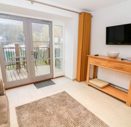 A living room with glass doors opening to a balcony with metal chairs and table a wooden console table below a mounted TV with a remote and bowl on it and a brown rug on the floor at Lowe-McConnell in Far Sawrey