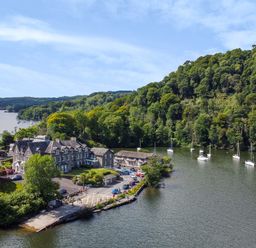 A waterfront estate with parked cars and moored boats next to a forested hillside at Lowe-McConnell in Far Sawrey
