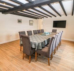 A dining room with a long table covered with a tablecloth and twelve chairs a wall-mounted television and wooden flooring at Trewen Botwnnog near Sarn Meyllteyrn