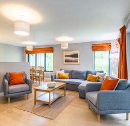 A living room with grey sofas and armchairs with orange cushions and curtains a wooden coffee table and dining table with chairs at Red Deer Lodge in Feshiebridge near Aviemore