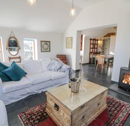 A living room with a white sofa and cushions a wooden chest used as a coffee table with a champagne bucket and glasses a fireplace and a dining area visible through an archway at Cwch Gwenyn in Llanfachraeth