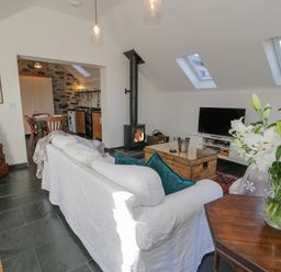 A living room with a white sofa and teal cushions a wood-burning stove a wooden coffee table and television next to a kitchen with wooden cabinets and dining table at Cwch Gwenyn in Llanfachraeth