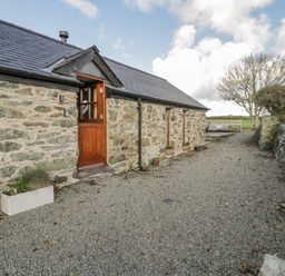 A stone cottage with a wooden door and two windows next to a gravel path with a seating area in a rural setting at Cwch Gwenyn in Llanfachraeth