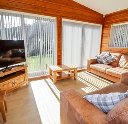 A living room with two brown sofas a wooden coffee table a flat screen tv and large windows with vertical blinds at Watersview Lodge Rudyard near Leek