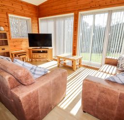 A living room with two brown sofas a wooden coffee table a TV on a wooden stand and large windows with vertical blinds at Watersview Lodge Rudyard near Leek