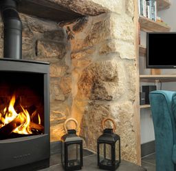 A wood burning stove with a fire inside stone walls two black lanterns a television and a blue chair at Stargazy Cottage in Praa Sands