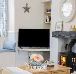 A living room with a wall-mounted TV window seat cushions a fireplace with a wood stove and a coffee table with flowers at Potter Hill Cottage in Pickering