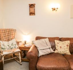 A corner of a living room with a leather sofa with cushions a wooden rocking chair with cushions and a blanket a side table with a lamp and a book on it at Beckfold Cottage in Broughton-In-Furness