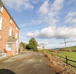 A brick house with multiple windows and a small porch next to an open paved area and fenced green fields at Squires Rest in Llanfair Caereinion