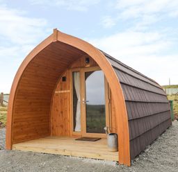 A small wooden pod with a curved roof gravel ground and a watering can outside at Garraidh Ghorm Pod in Conista