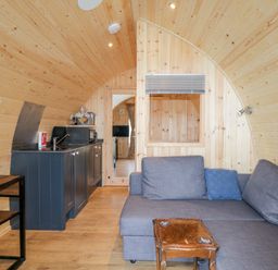 A small wooden pod interior with a kitchenette a grey sofa a wooden table and a doorway with a TV visible through it at Garraidh Ghorm Pod in Conista