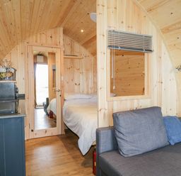 A small wooden room with a bed visible through a glass door a sofa with cushions and a kitchenette area at Garraidh Ghorm Pod in Conista